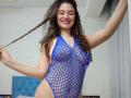 SanetPerlas with curly brown hair, smiling, wearing a blue fishnet bodysuit, exposing her tits and pussy, holding a braid to the side, in a bright room.