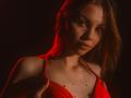 AgataRyili with red dress, small tits, and red nipple pasties, holding straps, red light, brown hair, serious expression, dark background.