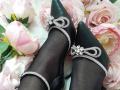 AmeliaMaiv's black pointed-toe shoes with silver diamond-encrusted bows and straps, covering her black-tan legs, set against pink roses and green leaves.