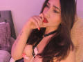 ElyRoss with dark hair, red lipstick, and black harness, biting finger, showing tits with pink nipple clamps, tattoo on left arm, purple lighting, bedroom setting.