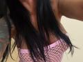 AndraD's black hair falls over her pink fishnet top, revealing her boobs. She has a tattoo on her left arm. The top has a white pattern.