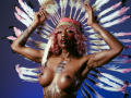 DaniaMarcell with pink hair, wearing a feathered headdress, topless, showing her big boobs, tattoos, and dark skin. Hands raised, blue background. Feathers in white, pink, and black.