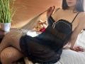 Tisia with long brown hair, wearing black lace lingerie, sheer mesh, and fishnet stockings, holding a feather, showing her tits and ass, sitting on a bed with a teddy.
