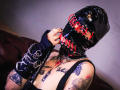 AmySpider wearing a shiny black latex mask with red eyelets, holding a black leather whip, sporting tattoos on her chest and arms, in a black tank top.