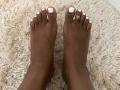 RoldanStefy's dark brown toes with white nail polish, wearing a gold ring on the right big toe, standing on a fluffy beige rug.