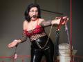 KarlaHartley with black hair, glasses, red-black fishnet top, black shiny pants, ball gag in mouth, holding leather and chain, pink cuffs, standing, looking forward, right arm extended.
