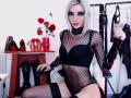 MisssDarkLady with blonde hair, black mesh lingerie, black bra, thigh-high stockings, holding black leather gloves, sitting on chair, red roses in vase, black boots, red heels.