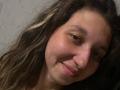 Sanella's face with wavy brown hair, light skin, and a slight smile. Her eyes are half-closed, and her nose is slightly upturned. She has a subtle, natural look.