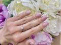 Olivias Hand mit rosafarben lackierten Nägeln, mit einem silbernen Ring mit einem kleinen Diamanten, liegt auf weißen und rosafarbenen Blumen. Ihre Haut ist hell, die Finger sind leicht gespreizt..
