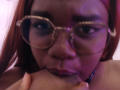 RoseBeckers with red hair, wearing glasses, sucking on a large, dark-skinned, flaccid, uncircumcised penis, holding it with her right hand, purple lighting in the background.