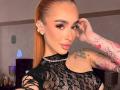 MarissaFex with orange ponytail, wearing black lace top, showing tattooed arm, star earrings, and pouty lips, touching her head. Boobs partially visible through lace.