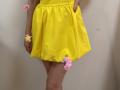 MiraMixi's yellow dress, hands with pink flowers, yellow ring, red nails, and a pink flower on her thigh, standing against a plain wall.