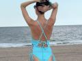 Nalini, in un costume intero blu, con le braccia alzate, mostra la schiena con spalline incrociate e tagli laterali, in piedi su una spiaggia con l'oceano sullo sfondo..
