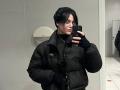 HanhFontillas, wearing a black puffy jacket, black gloves, and glasses, takes a mirror selfie with a black phone in a bathroom, showing short black hair and a neutral expression.