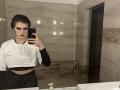 HanhFontillas with dark makeup, wearing a white crop top, black long sleeves, and black pants, taking a mirror selfie in a tiled bathroom. Hand on phone, serious expression.