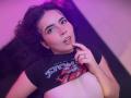 AliinaFontaine with curly black hair, raising her black "Stone Gargoyles" shirt to expose her small, pale tits, finger touching her parted lips, under pink and purple lighting.