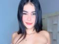 NahomiMillerr with black hair, pink cheeks, and red lipstick, smiling, topless, showing small tits, wearing a necklace with a tiny pendant, in a brightly lit room.