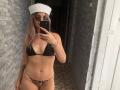 Milagres in a black bikini top and leopard print thong, wearing a white sailor hat and glasses, taking a mirror selfie with a phone, showing her tits and pussy.