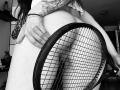 SujannaVarma's tattooed hand grips a tennis racket against her bare ass, showing a bracelet and ring. Black and white image, SujannaVarma's long hair partially visible.