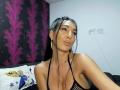 BrunetteBabe69 mit dunklem Haar, trägt ein schwarzes, durchsichtiges Top mit Halter, zeigt ihre großen Brüste, volle Lippen und goldene Ohrringe, in einem Schlafzimmer. Ihre Haut ist gebräunt und sie schaut in die Kamera..