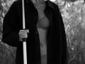 Black-and-white photo of EolantaMarina with blonde hair, wearing an open dark jacket, exposing her bra-covered tits and underwear, holding a walking stick in her right hand.