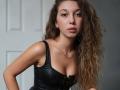 Sanella with wavy brown hair, wearing a black leather top that shows her small tits, has a neutral expression, and is standing against a white door.