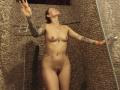 SarahRousen, naked with tattoos, stands in a pebble-textured shower, arms raised, eyes closed, hands on glass, water droplets on her skin, small tits, shaved pussy, red wristband.