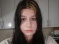 AlicaMihal has pale skin, long black hair with a blonde streak, brown eyes, and pink lips, wearing a gray shirt, looking straight at the camera with a neutral expression.