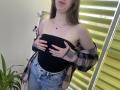 Nesie with hands covering her black strapless top, wearing a pink plaid shirt off her shoulders and blue jeans. Her tits are visible. She's standing by yellow blinds.