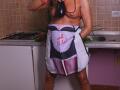 KarlaHartley stands in a kitchen, wearing a black and white maid apron, exposing her tits and holding a purple eggplant to her mouth, with a red bead necklace.