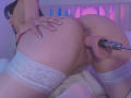 ArianaRogers's ass is plugged with a pink butt plug, wearing white lace thigh-highs, hand on her butt, purple and pink lighting, visible anus and labia, long nails.