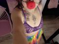 ArianaBlonde with red ball gag, purple lace bra, colorful tie-dye bottoms, tattoos on chest and neck, holding camera, wearing glasses, wooden floor, pink chair.