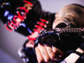AmySpider in black latex with red and black leather bondage mask, holding cuffs with a chain, focusing on her hand. Tattoo visible on her arm. Intense BDSM scene.
