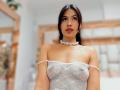 MelisaDaltonn with dark brown hair, wearing a white, off-shoulder, mesh top that shows her small, perky tits and a white lace choker, standing in a bright room.