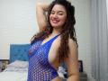 SanetPerlas with curly brown hair, smiling, wearing a blue fishnet top that exposes her large, round boobs and side-ass, standing in a bedroom.