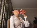 KassandraDalal with glasses, drinking from a pink bottle, wearing a white shirt that shows her tits, sitting in a chair, dark curtains behind her.