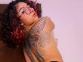 Topless KanelaMiller with curly red-tinted hair, glasses, and large butterfly tattoo on her back, looking over shoulder, showing small tits and a nipple piercing.