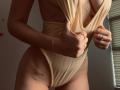 SugarrX in a beige one-piece, pulling down the top to reveal small tits with tattoos on her side and lower belly, wearing a gold bracelet and rings.
