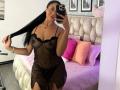 CassieFox with long black hair, wearing a sheer black lace dress, showing her tits and black panties, holding her hair, smiling, taking a mirror selfie in a bedroom.