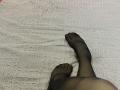 Les jambes noires et transparentes de Destinis, avec ses pieds, reposent sur une couverture blanche texturée. Ses orteils sont légèrement écartés, révélant la texture du tissu. Ses jambes sont croisées aux chevilles..