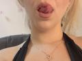 SugarrX's face with tongue out, pink glossy lips, fair skin, black halter top, small gold necklace with heart pendant, blonde hair, slightly visible earrings, and a slight double chin.