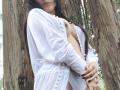 TaniaSandersX with large tits, wearing a white, sheer, button-up, exposing one boob, standing against a giant tree, long brown hair, dark nails, and a ring.