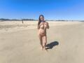 DosCalientes standing on a sandy beach, topless with hands covering her tits, wearing orange bikini bottoms, long black hair, blue sky, barefoot, sunny, clear shadow on sand.