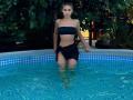 MilagrosEdmisten stands in a blue-tiled pool, water up to her thighs, wearing a black strapless top and black shorts, her hand on the pool's edge, intense gaze, dark hair slicked back.