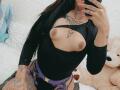 DenyChantal with exposed tits, black zip-up top, purple strappy belt, tattoos, long black hair, pink nails, holding phone, sitting on white bed with teddy bear.