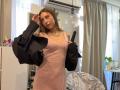ErinMarrujo trägt ein pinkes Kleid und hält eine schwarze Jacke. Ihre Brüste sind durch das Kleid sichtbar. Sie hat langes, braunes Haar und trägt eine Halskette. Sie steht in einem Raum mit einem Stuhl und hängenden Lampen..