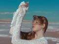 StellaLane with her eyes closed, wearing a white lace long-sleeve top, arms raised, brown hair blowing, subtle makeup, small hoop earrings, clear blue sky, calm expression.