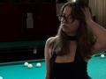 LeScharwath with glasses, black choker, and black dress, hand in brown hair, standing by green pool table, looking serious, with white pool balls in the background.