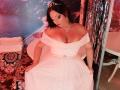 AmberSunshine con grandi seni, vestita con un abito da sposa bianco a spalle scoperte, che tiene la gonna, indossando una tiara, una collana e rossetto rosso, in piedi in una stanza con festoni argentati..