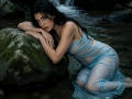 ZoeeMar con un abito blu trasparente e bagnato, inginocchiata vicino a una roccia nel ruscello, che guarda la fotocamera con un'espressione seducente, con lunghi capelli neri, piccoli seni e pelle chiara..